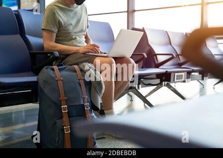 uomo asiatico viaggiatore d'aria maschile con maschera nera seduta in un'area di attesa nell'edificio del terminal dell'aeroporto utilizzando un computer portatile Foto Stock