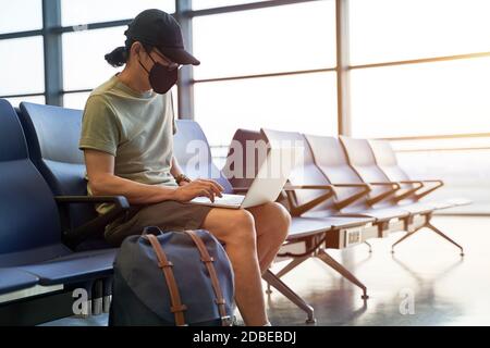 uomo asiatico viaggiatore d'aria maschile con maschera nera seduta nell'area d'attesa emply nell'edificio del terminal dell'aeroporto usando il laptop computer Foto Stock