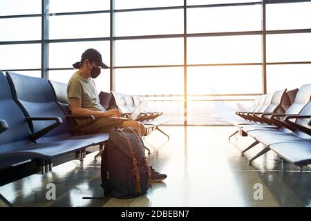 uomo asiatico viaggiatore d'aria maschile con maschera nera seduta nell'area d'attesa emply nell'edificio del terminal dell'aeroporto usando il laptop computer Foto Stock