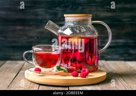 Tè alle erbe con frutti di bosco, lamponi, foglie di menta e fiori di ibisco in teiera di vetro e tazza su tavola di legno Medicina per la vitamina fredda bevanda rustica Foto Stock