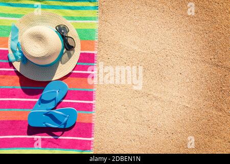 Telo da spiaggia con cappello, occhiali da sole e infradito fotografato dall'alto sulla spiaggia di sabbia, caldi accessori estivi, destinazione di vacanza, soleggiata spiaggia tropicale sfondo Foto Stock