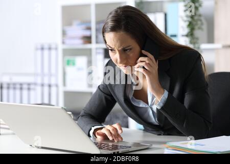 Donna dirigente sospetta che chiama con uno smartphone guardando il computer portatile seduto sulla scrivania in ufficio Foto Stock