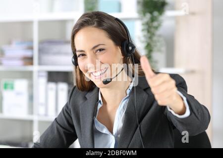 Donna di telemarketer felice con i pollici in su che indossa la cuffia che guarda la macchina fotografica seduto all'ufficio Foto Stock