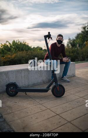 Giovane uomo su uno skateboard elettrico che usa un telefono cellulare seduto su una panchina con il suo casco. Trasporto alternativo. Foto Stock