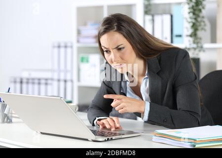 Donna dirigente sospetta che legge e punta sul computer portatile seduto su una scrivania in ufficio Foto Stock