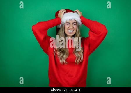 Giovane bella ragazza che indossa il cappello di Natale di Santa tenuta regalo sopra sfondo verde isolato che soffre di mal di testa disperato e stressato perché pa Foto Stock