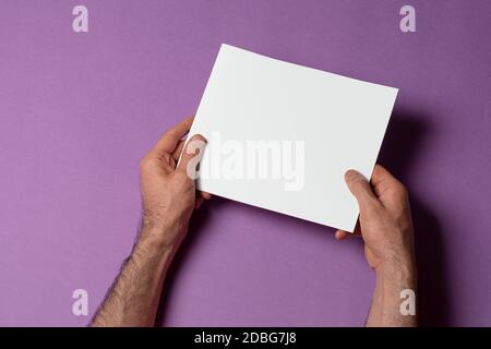 Mani maschili con un catalogo di proporzioni quadrate con copertina vuota su sfondo viola, modello di serie mock-up pronto per il tuo percorso di selezione del design inc Foto Stock