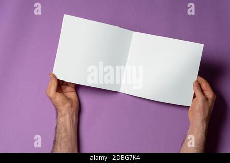 Mani maschili che tengono un catalogo aperto con pagine vuote su sfondo viola, modello di serie mock-up pronto per il percorso di selezione del progetto incluso Foto Stock