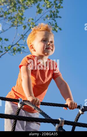 Piccolo ragazzo in piedi su corde in un parco giochi e guardando con sicurezza in lontananza. Una nuova piattaforma in crescita... Foto Stock