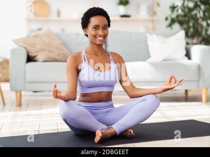 Donna nera sorridente in abbigliamento sportivo seduto sul tappetino yoga in posa lotus e meditando o facendo esercizi di respirazione, in interni Foto Stock