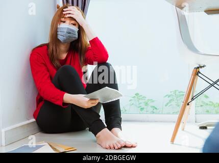 Asian giovane donna triste abbracciare il suo ginocchio e gridare indossando maschera facciale protettiva disoccupato quarantine malattia coronavirus o COVID-19 sua seduta da sola su fl Foto Stock