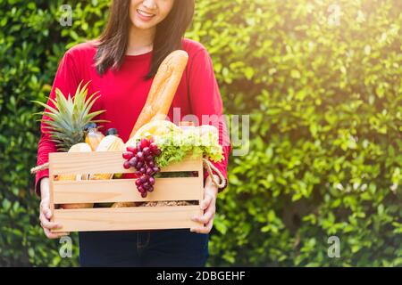 Ritratto di Asian bella giovane donna contadina in piedi sorride e tenendo pieno cibo fresco frutta verdura cruda in una scatola di legno nelle sue mani sul verde Foto Stock