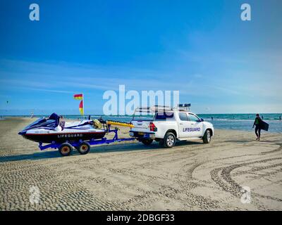 Un giovane body boarder cammina verso il surf mentre i bagnini forniscono sicurezza in un camion fresco e moto d'acqua nel rimorchio. Foto Stock