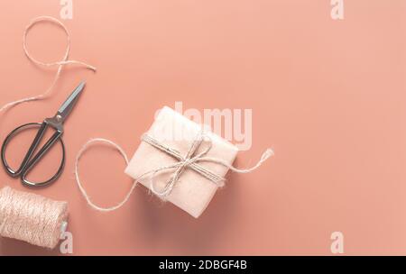 Confezione regalo in carta artigianale su sfondo color pesca, forbici e carta da imballaggio nelle vicinanze. Il concetto di preparare i regali per la festa. Vista dall'alto, spazio di copia Foto Stock