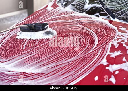 Cappuccio anteriore auto rosso lavato in autolavaggio, dettaglio sulla spazzola lasciando colpi in schiuma di sapone bianco. Foto Stock