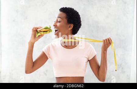 Disgregazione della dieta. Signora nera cercando di mangiare hamburger malsano, tirandosi via da metro nastro, fondo grigio Foto Stock