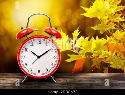 Orologio rosso vintage con sveglia sullo sfondo di foglie di acero giallo autunno. Ritorno all'inverno. Il vecchio orologio accanto all'autunno parte su un ta di legno Foto Stock