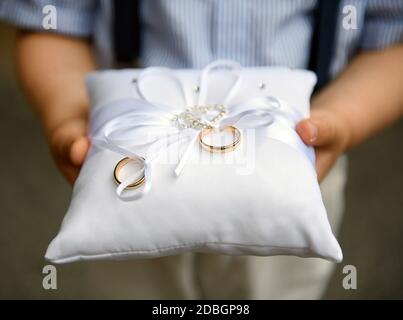 Pageboy che tiene due bande o anelli di nozze d'oro su un cuscino decorato con arco bianco durante la cerimonia di matrimonio a. una chiesa in primo piano Foto Stock