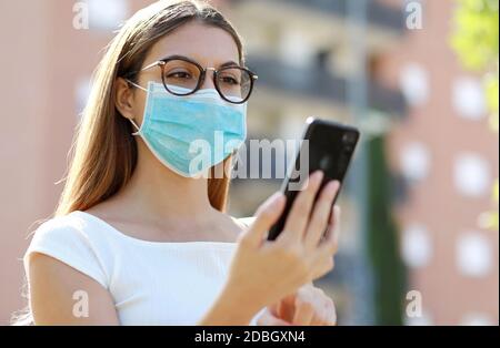 Primo piano di giovane donna con maschera medica che digita su smartphone in città strada. Piuttosto casuale ragazza lettura messaggio su mobile chattare app outdoor. Foto Stock