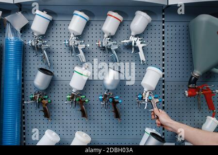 Il lavoratore maschio in uniforme tiene la pistola a spruzzo pneumatica della vernice nel deposito attrezzi. Scelta di attrezzature professionali nel negozio di ferramenta, supermercato di strumenti Foto Stock