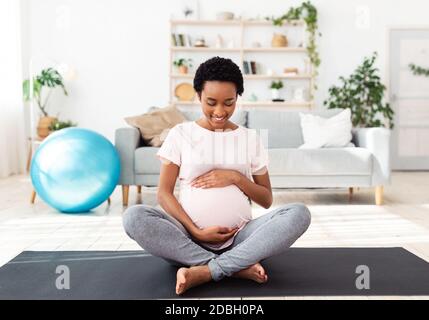 Concetto di sport e gravidanza. Bella donna nera in attesa abbracciare il suo grande ventre mentre medita sul tappetino a casa Foto Stock