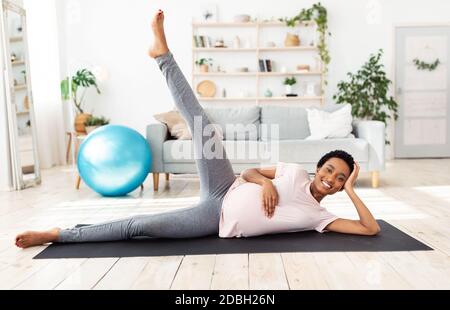 Ritratto a lunghezza intera di donna nera in attesa che si esercita sul tappetino yoga, sollevando la gamba, sentendosi sano a casa Foto Stock