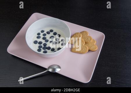 Colazione. Yogurt con biscotti a forma di cuore ai mirtilli. Ingredienti in un vassoio rosa su un tavolo di legno nero. Foto Stock