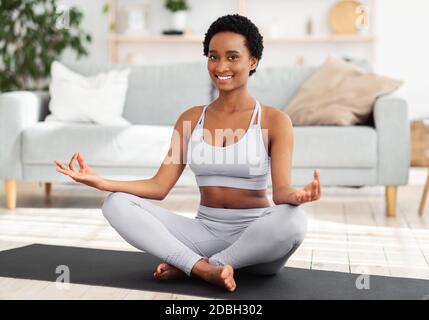 Sport domestici durante il concetto di blocco. Donna nera positiva meditando in lotus posa a casa Foto Stock