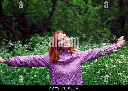 Donna rossa felice con mani sparse in piedi in una foresta con occhi chiusi e sorriso gioioso. Foto Stock