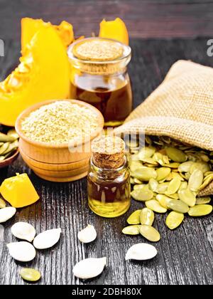 Olio di zucca in un flaconcino di vetro e un vaso, farina in un recipiente, semi in un cucchiaio e sacchetto, fette di verdure su sfondo di legno scuro Foto Stock