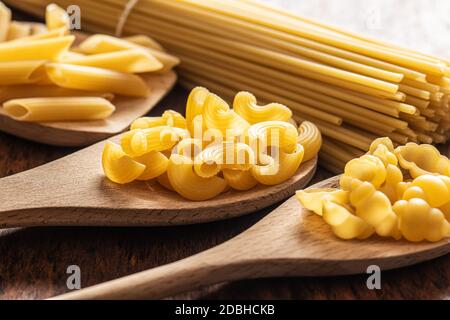 Vari tipi di pasta italiana cruda in cucchiai di legno su tavola di legno. Foto Stock