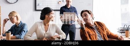 Felici colleghi multiculturali che parlano mentre si siedono ai tavoli in ufficio con i colleghi sullo sfondo, banner Foto Stock
