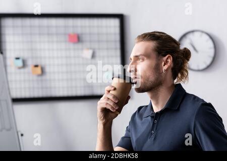 Lavoratore sicuro che tiene la tazza di carta e guarda via con organizer mesh sfocato e orologio sullo sfondo Foto Stock