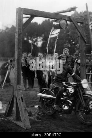 Membro del Motor HJ durante un test di abilità di guida durante il festival sportivo della gioventù Hitler al Westend Sportplatz di Berlino. Guida sotto un secchio d'acqua. Il pubblico è in background. Sulla destra della foto sono presenti altri membri dell'HJ che guardano la scena. Guida una moto del marchio DKW tipo SB 350 fuoristrada. Sullo sfondo è rimasto un fotografo con una fotocamera di grande formato. Foto Stock