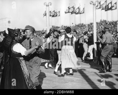 Uomini in uniforme e costume tradizionale ballano in allegria con donne in costume tradizionale su un podio al festival pubblico sul campo Zeppelin. Foto Stock