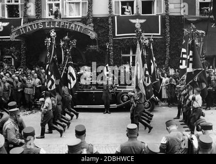 I membri della SA marciano oltre Adolf Hitler con le loro bandiere di tempesta. Rudolf Hess è in piedi davanti alla macchina. Dietro l'auto di Adolf Hitler c'è la Bandiera di sangue (Blutfahne). Sullo sfondo si trova l'ingresso alla Haus Elephant. Foto Stock
