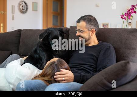 Giovane coppia felice sul divano a casa con il loro cane nero. Donna che riposa la testa sulle gambe del suo compagno. Foto Stock
