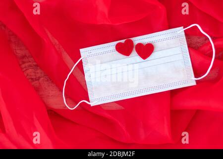 Maschera medica protettiva blu con due cuori rossi su sfondo rosso. Regalo romantico per San Valentino. Amore. Concetto di proposta di matrimonio. Messa a fuoco morbida. Spazio di copia. Vista dall'alto Foto Stock