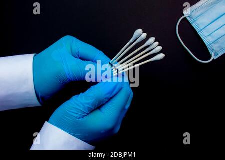 Un medico personale che tiene i tamponi della gola accanto a una maschera medica viso su uno sfondo nero. Foto Stock