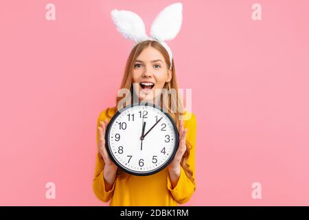 Buona Pasqua. Bella ragazza in Pasqua Bunny orecchie tenendo un orologio da parete in mano, su uno sfondo rosa isolato Foto Stock