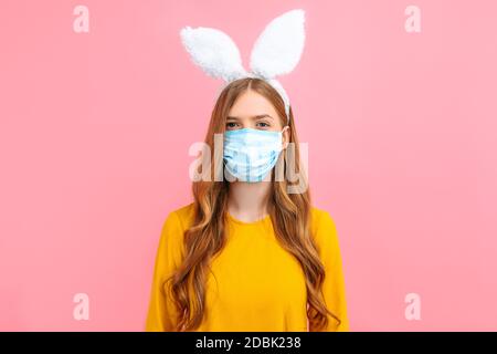 Una giovane ragazza nelle orecchie di un coniglietto pasquale, indossando una maschera medica sul viso contro le malattie virali, su uno sfondo rosa isolato Foto Stock