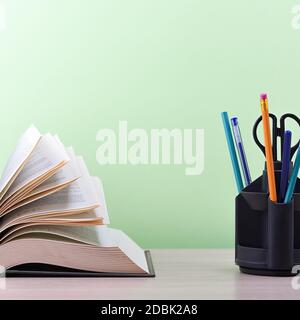 Un libro e un supporto con penne, matite e forbici su un tavolo di legno chiaro. Il libro è aperto, le foglie della pagina sono sbiadite, uno sfondo verde Foto Stock