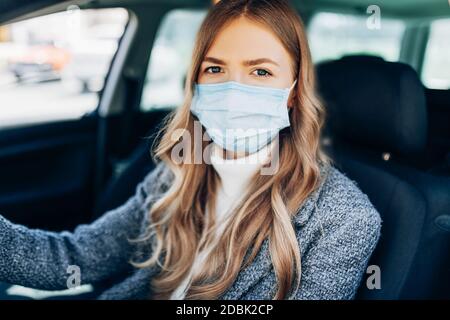 Bella ragazza giovane in una maschera seduta in un'auto, maschera protettiva contro il coronavirus, conducente su una strada della città durante un focolaio di coronavirus, covid-19 Foto Stock