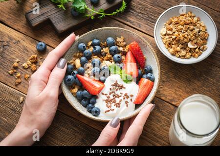 Recipiente di granola con frutti di bosco e yogurt in mani femminili su fondo di legno. Cibo pulito, concetto di dieta Foto Stock