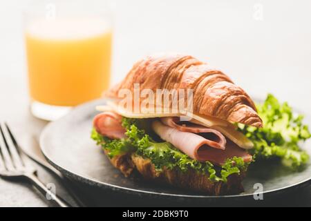 Panino di croissant con prosciutto, formaggio e insalata di lattuga serviti con un bicchiere di succo d'arancia. Messa a fuoco selettiva, immagine a toni Foto Stock