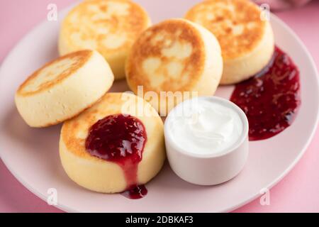 Frittelle di Syrniki o formaggio con marmellata di lamponi e panna acida su un piatto rosa, vista closeup Foto Stock