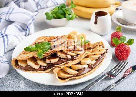 Crepes farcito con crema di nocciole al cioccolato e fette di banana su piatto bianco. Dessert crepes francese o blini russo Foto Stock