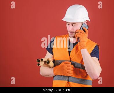 Ingegnere, architetto, costruttore sul volto occupato parla sullo smartphone mentre tiene progetti. Uomo, caposquadra in casco, supervisiona la costruzione sul telefono, sfondo rosso. Concetto di architetto. Foto Stock