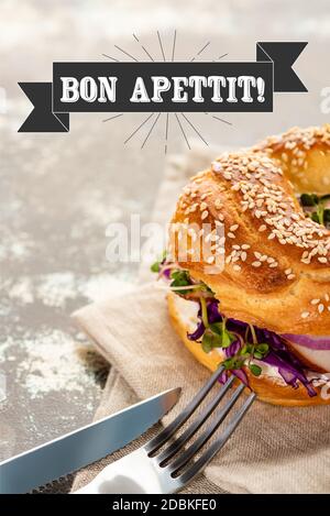 bagel fresco con carne su tovagliolo con posate vicino al bon scritta apettita su superficie testurizzata Foto Stock