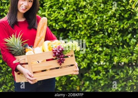 Ritratto di Asian bella giovane donna contadina in piedi sorride e tenendo pieno cibo fresco frutta verdura cruda in una scatola di legno nelle sue mani sul verde Foto Stock